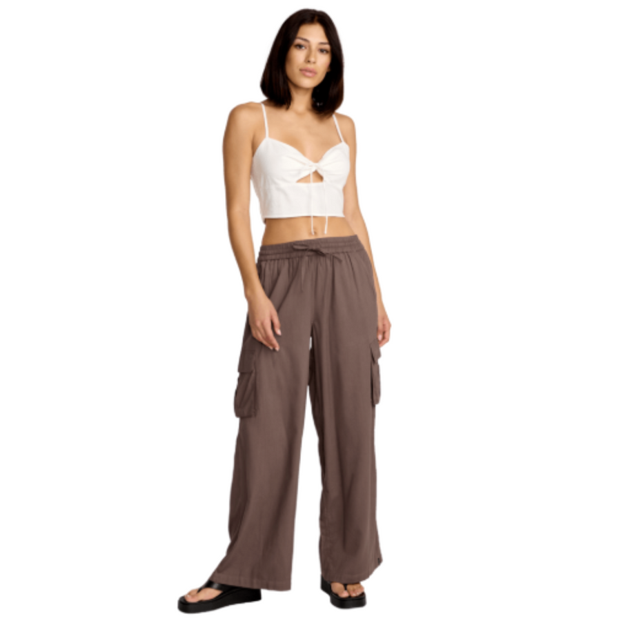 Woman wearing a white crop top and brown wide-leg volcom lived in beach pants on a white background