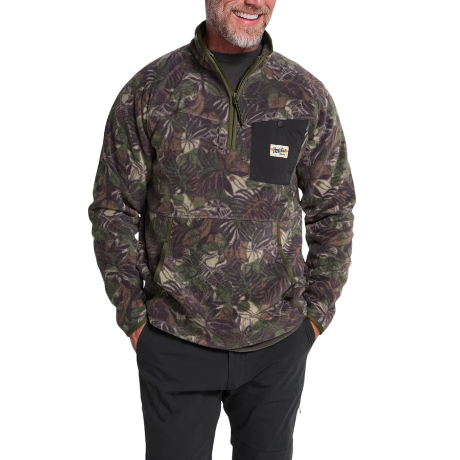 A long-sleeve quarter-zip howler brothers pullover fleece, featuring a dark green and brown tropical leaf camouflage pattern. It has a black chest pocket with a white and red Howler logo patch, a stand-up collar, and zippered hand pockets.