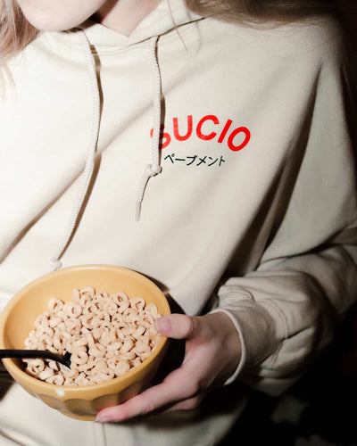 Person wearing a beige hoodie with 'SUCIO' branding, holding a bowl of cereal.