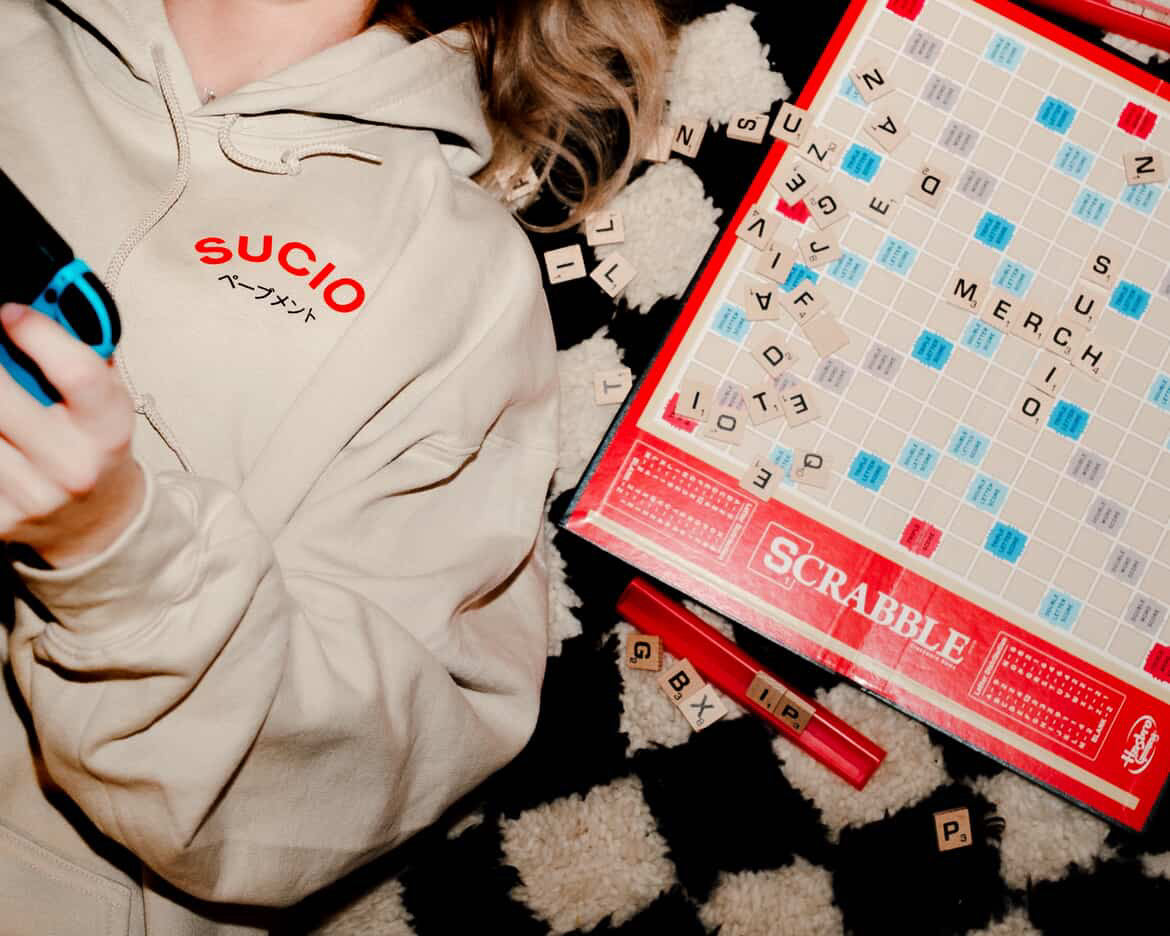 Person wearing a beige hoodie with 'SUCIO' text, holding a phone, next to a Scrabble board game.