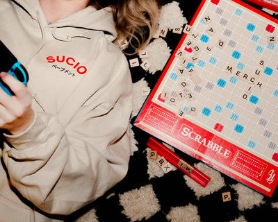 Person wearing a beige hoodie with 'SUCIO' text, holding a phone, next to a Scrabble board game.