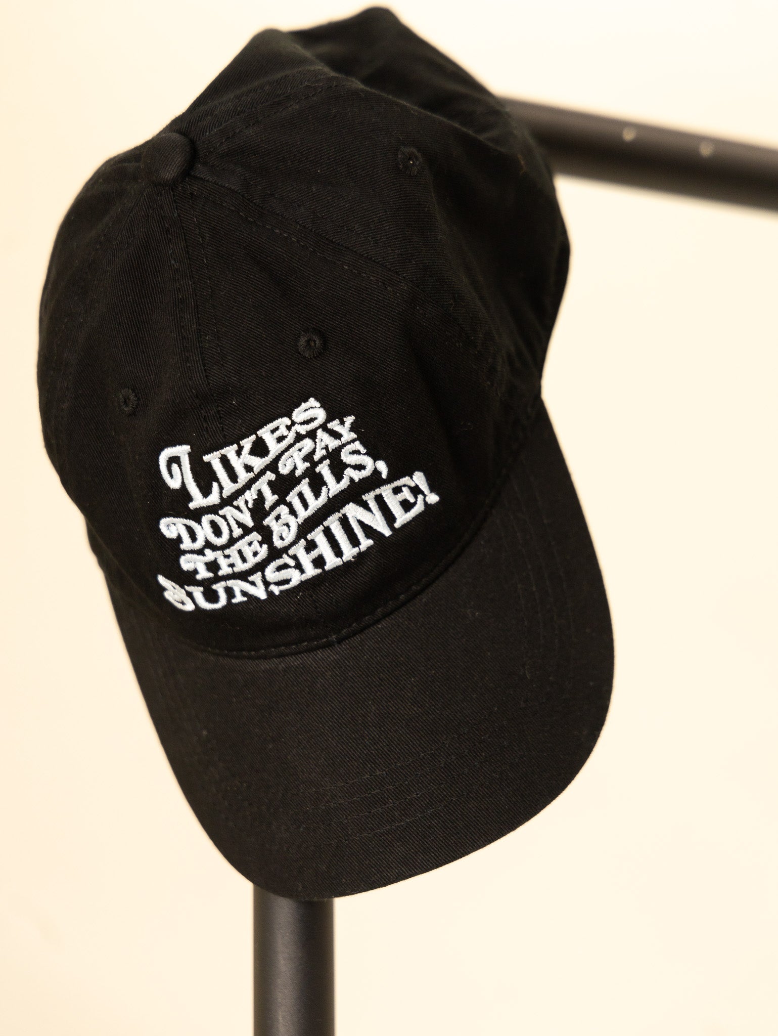 black embroidered hat with the phrase likes don't pay the bills sunshine