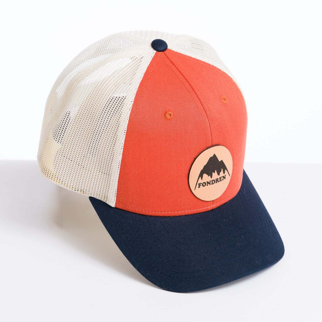 Trucker hat with navy bill, orange front panel and white back with a leather circle patch with the words fondren and a picture of a mountain