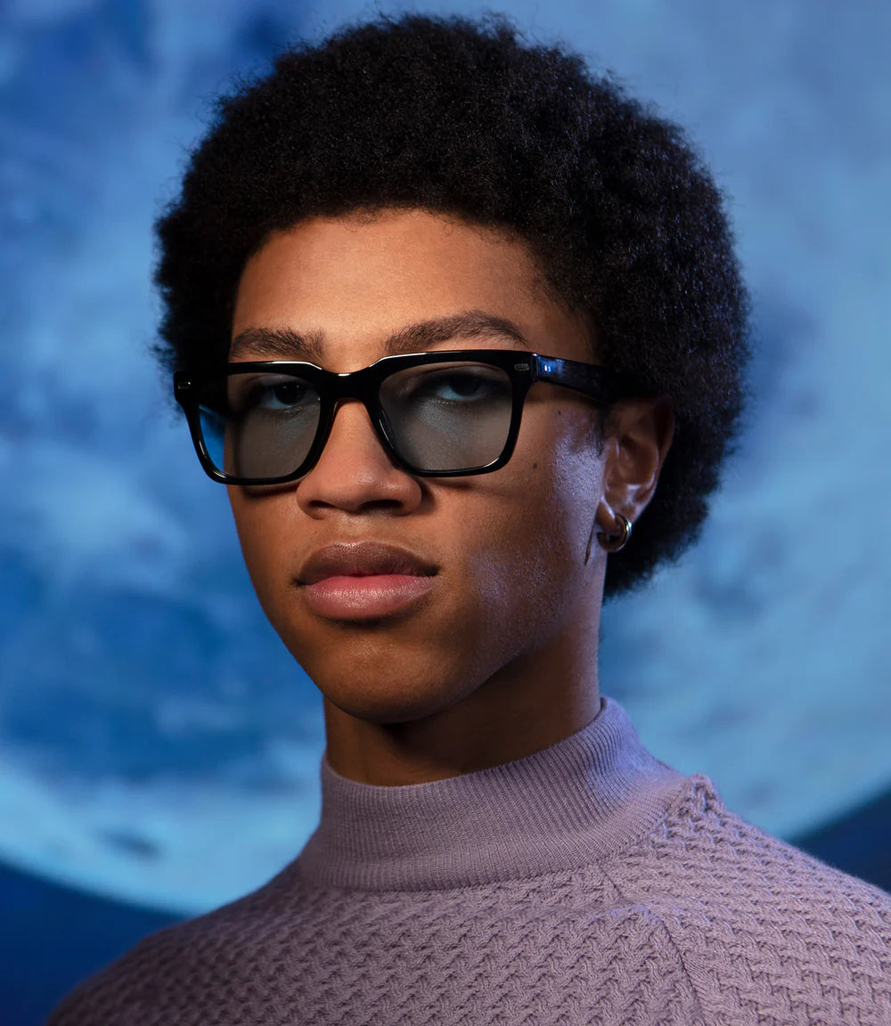 Person wearing black spitfire sunglasses and a light purple sweater against a blue background