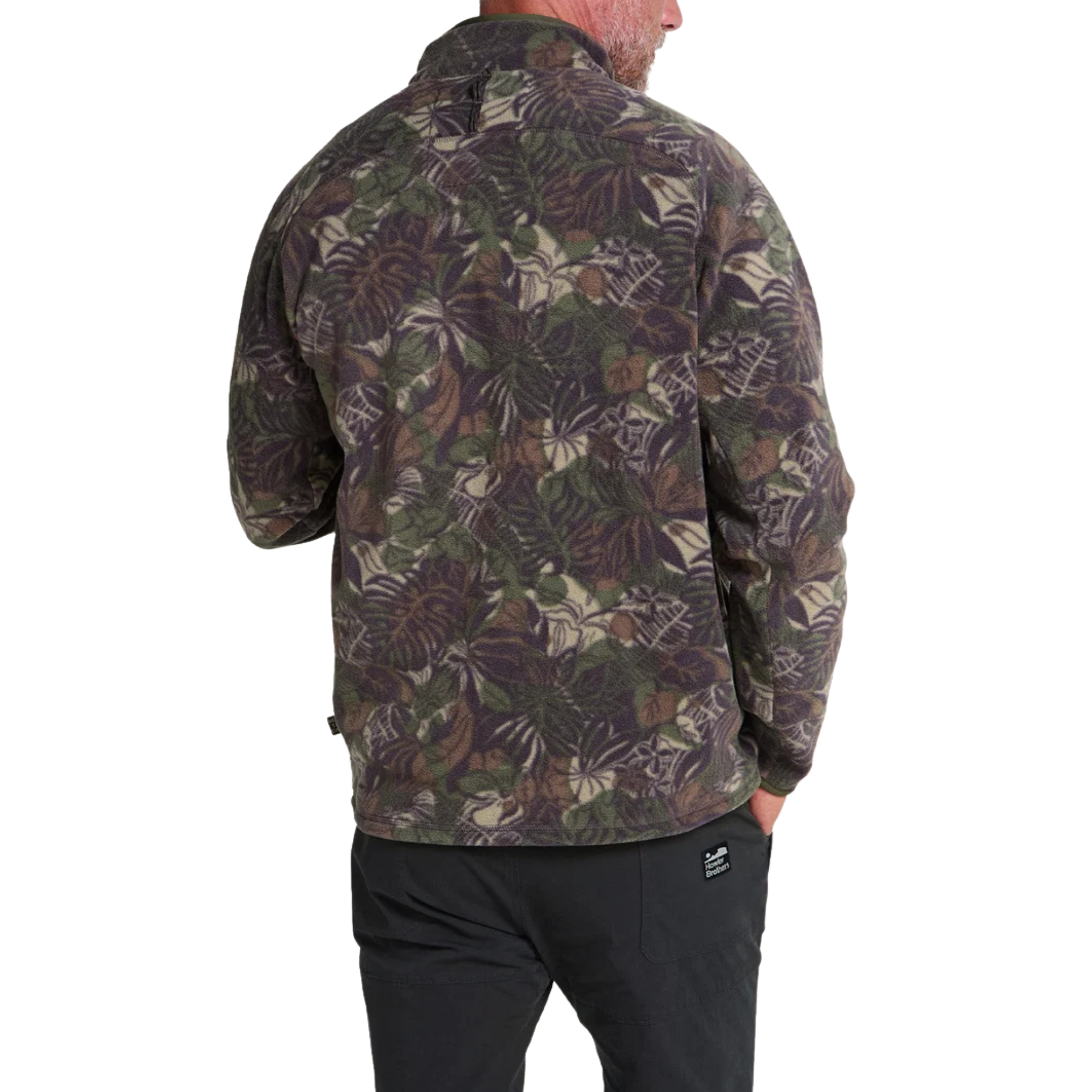 a man wearing A long-sleeve quarter-zip howler brothers pullover fleece, featuring a dark green and brown tropical leaf camouflage pattern. It has a black chest pocket with a white and red Howler logo patch, a stand-up collar, and zippered hand pockets.