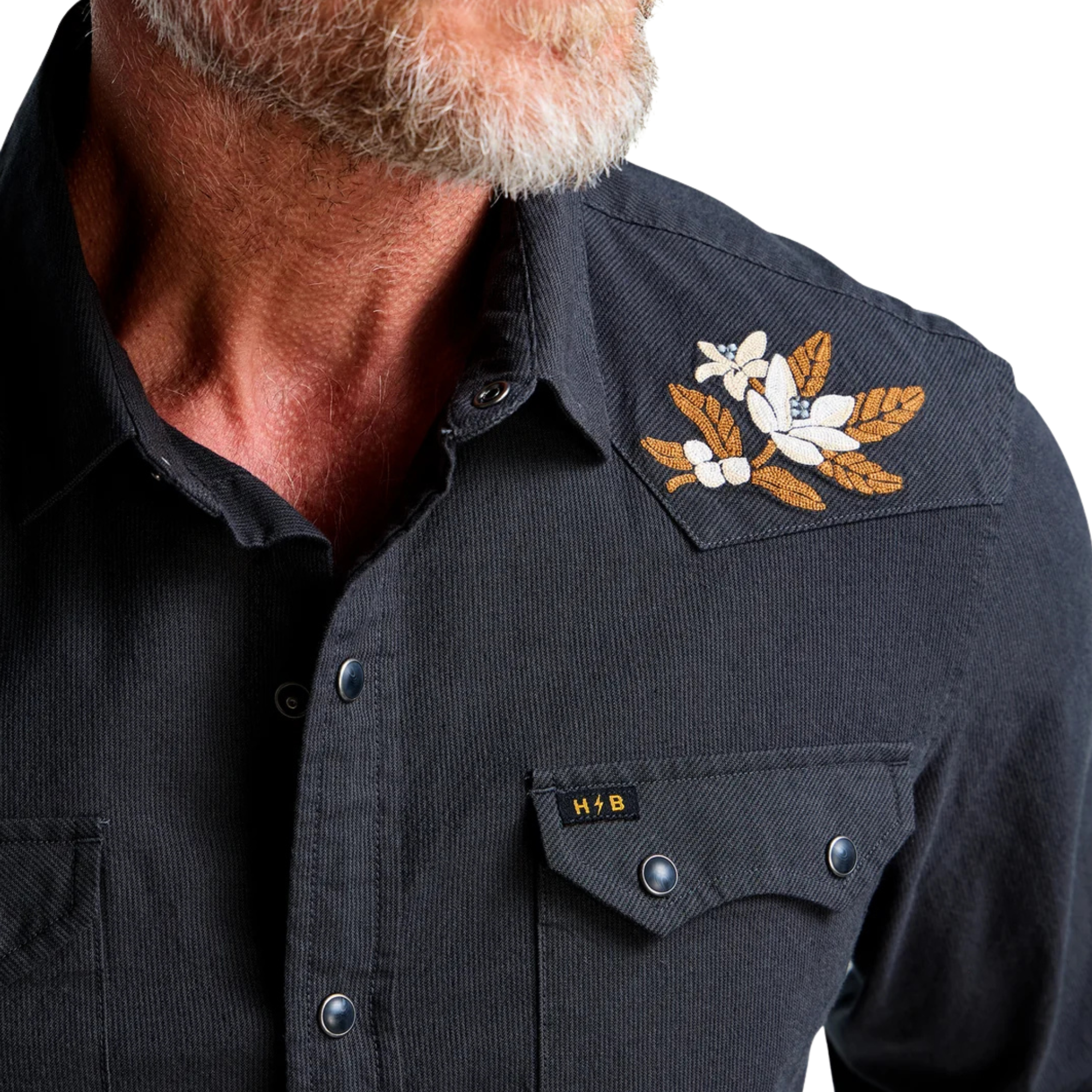 close up of a man wearing a howler dark navy western-style button-up shirt with embroidered eagle details on the shoulders and beige pants, photographed against a plain white background.