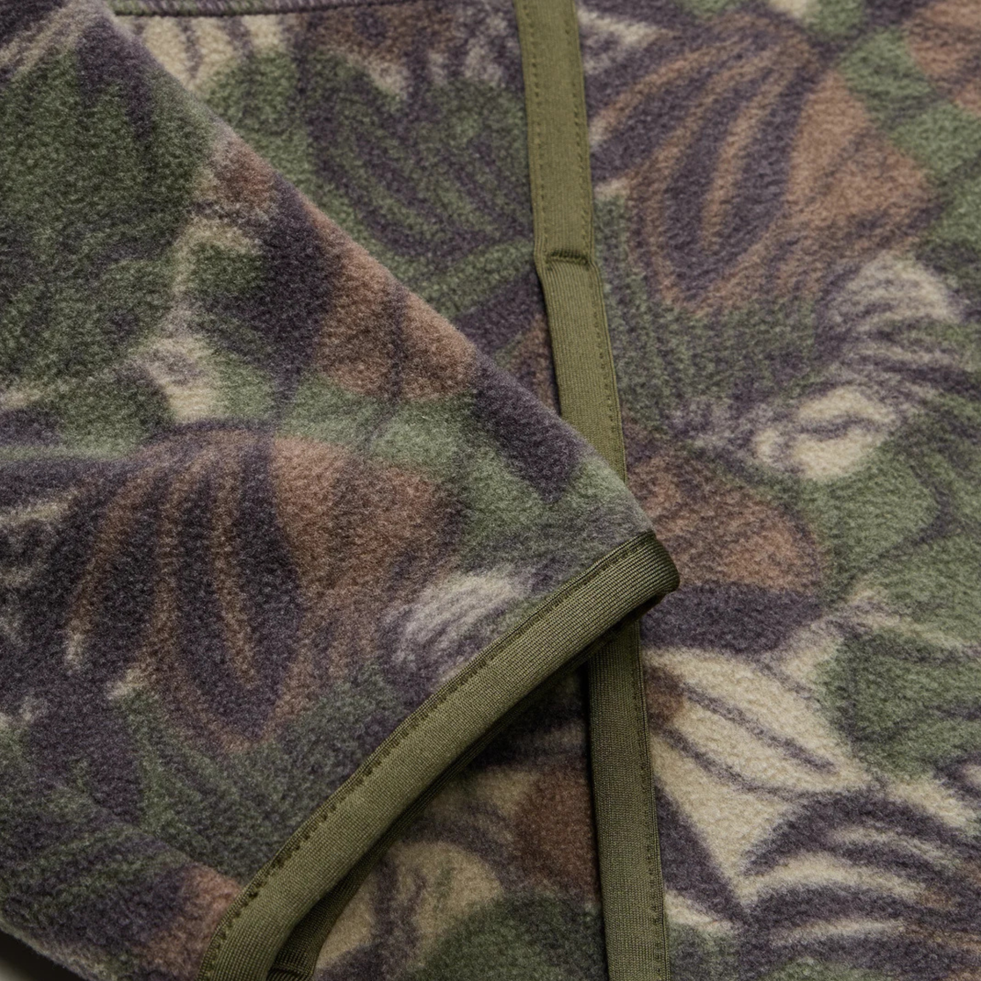 close up of A long-sleeve quarter-zip howler brothers pullover fleece, featuring a dark green and brown tropical leaf camouflage pattern. It has a black chest pocket with a white and red Howler logo patch, a stand-up collar, and zippered hand pockets.