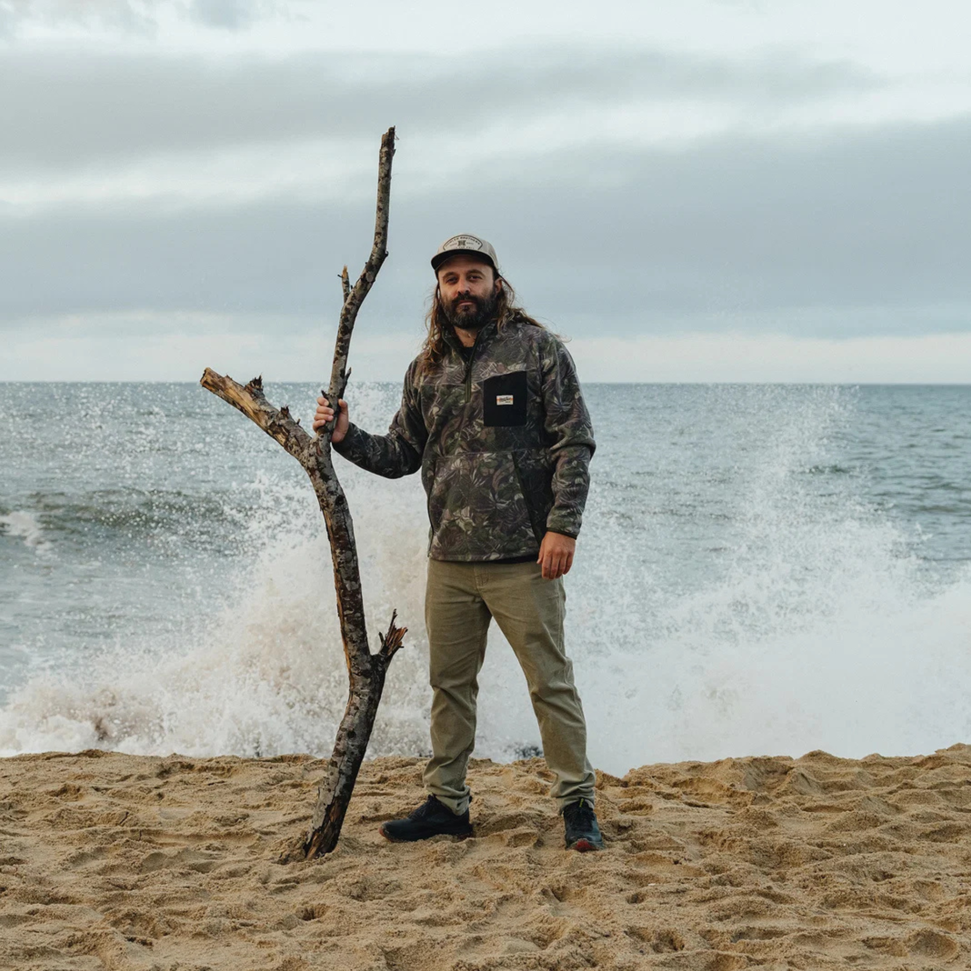 A man stands on a sandy beach holding a large bare tree branch, wearing a camouflage hooded jacket, olive pants, and a cap, with ocean waves crashing behind him under an overcast sky.