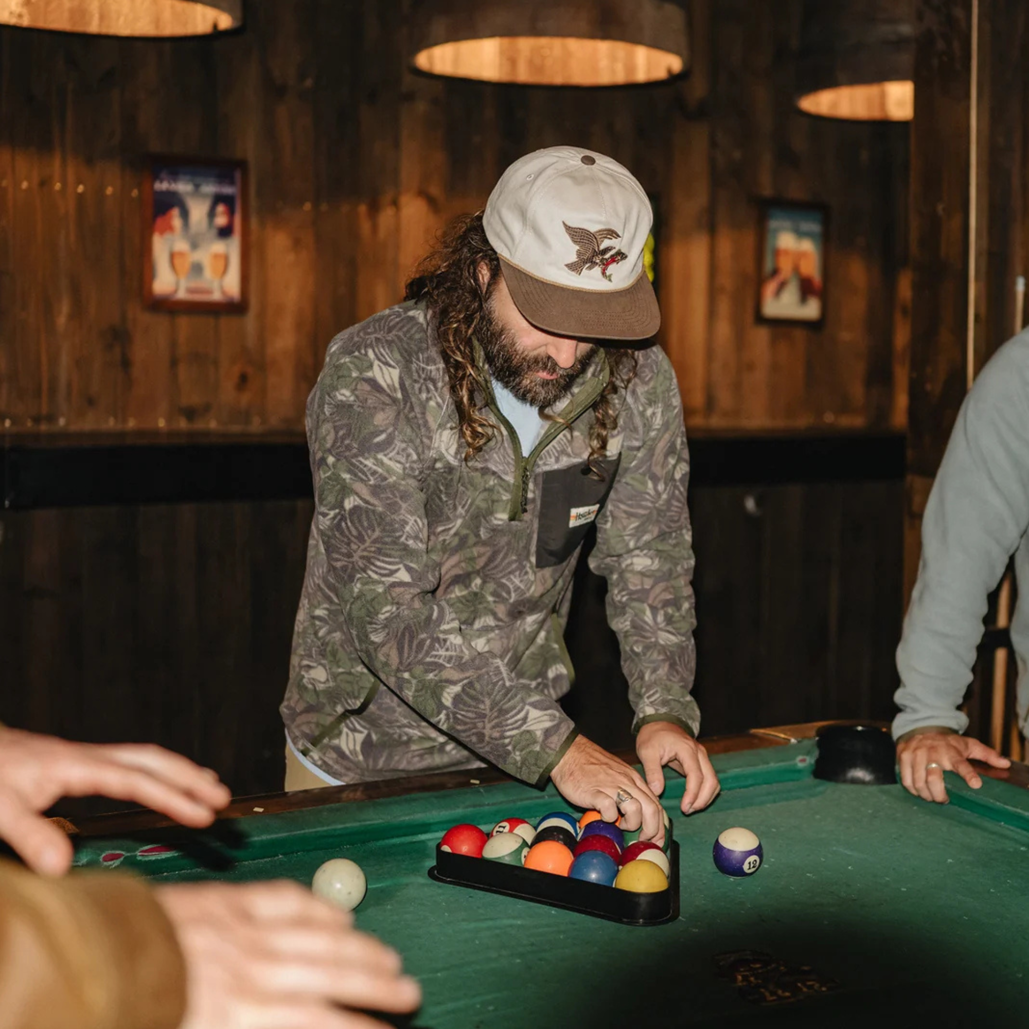 A man in a camouflage fleece pullover, beige baseball cap, and face mask leans over a green pool table, selecting a billiard ball. The setting is a dimly lit bar with wooden walls and hanging lights.