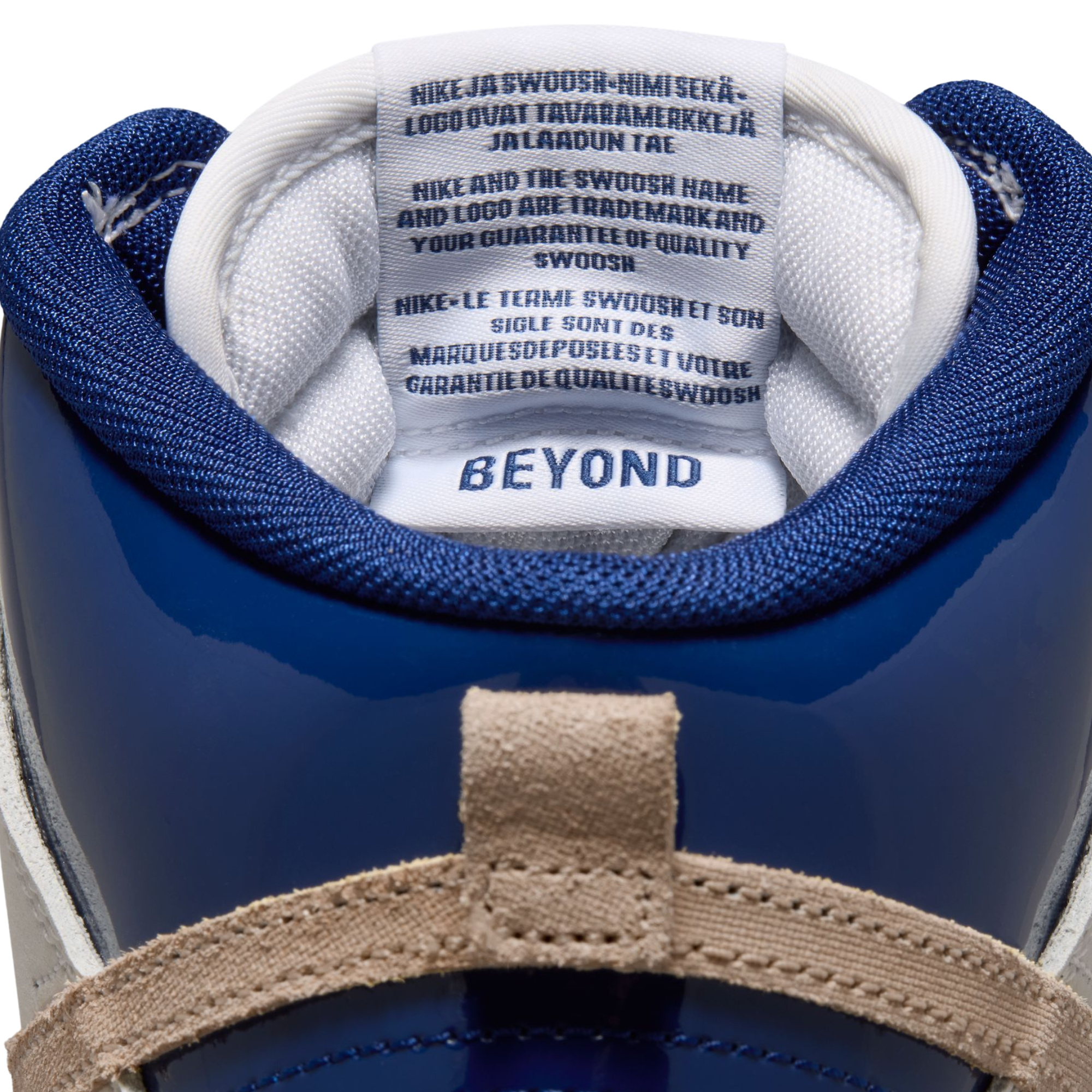 Close-up of the collar of a blue and white Nike sb dunk high shoe, showing the interior tag with 'BEYOND' branding and multilingual quality text, plus a beige suede strap at the top.