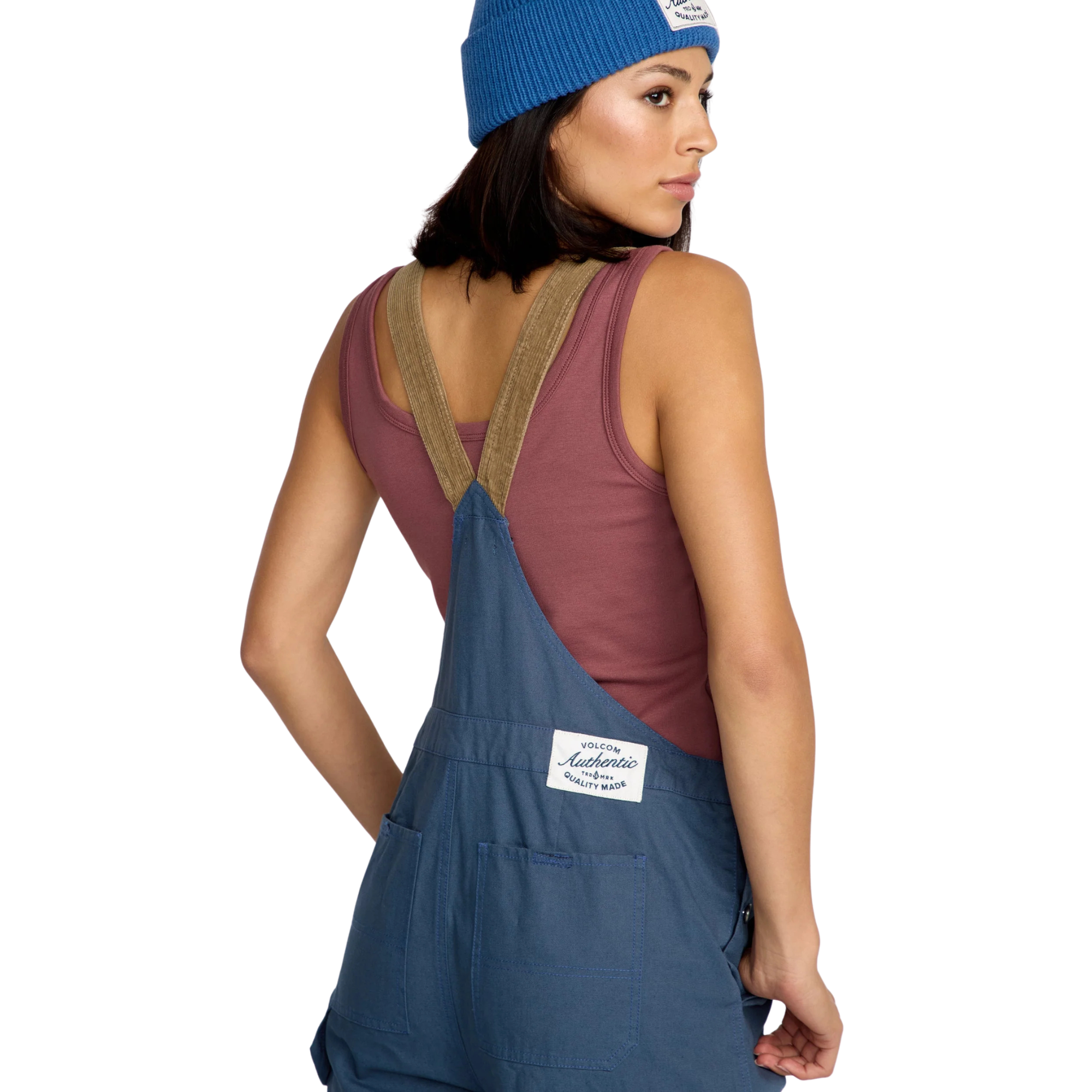 Woman wearing blue volcom full cut overalls with brown cordoroy straps and a matching beanie on a white background