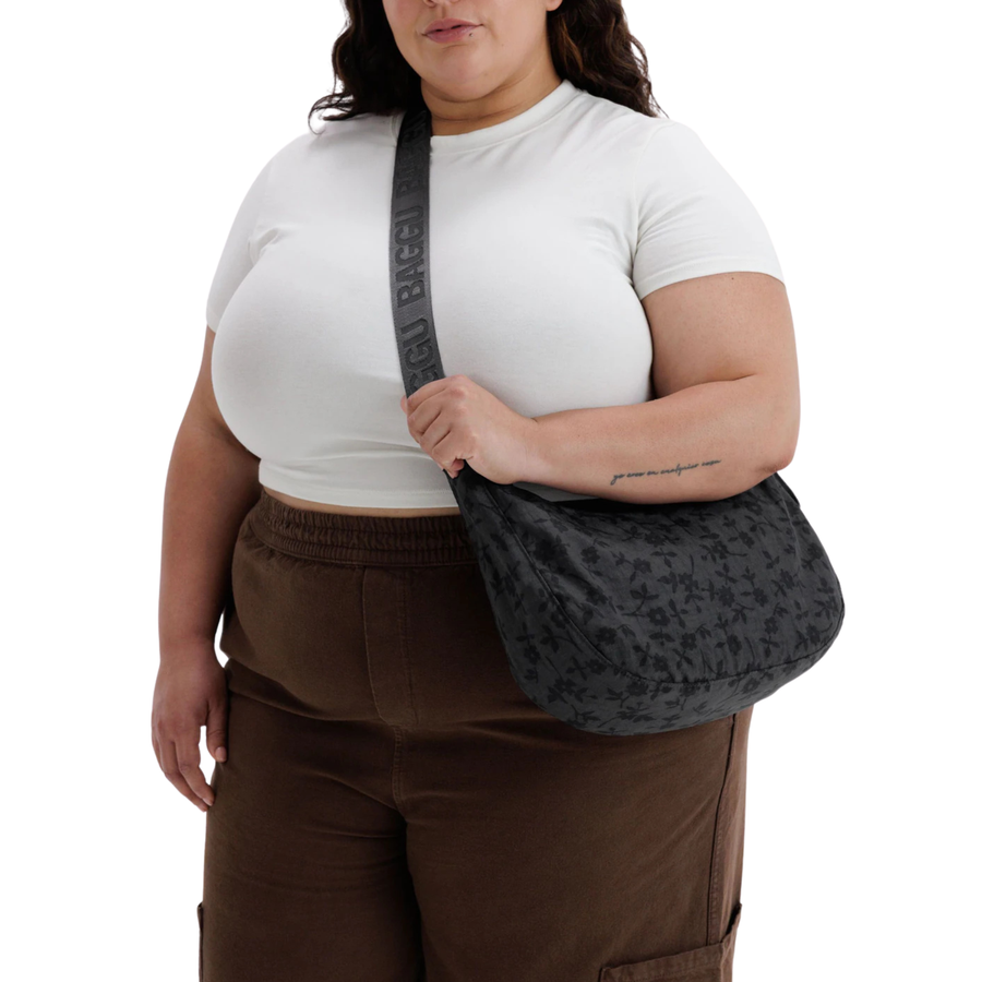 A black crescent-shaped baggu shoulder bag with a subtle tonal floral pattern, featuring a matching black strap with embossed lettering, shown against a plain white background.