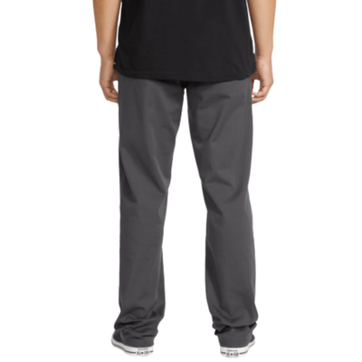 A person wearing dark gray volcom frickin stretch chino pants straight-leg work pants, a black t-shirt, and black canvas sneakers, shown from the waist down against a plain white background.