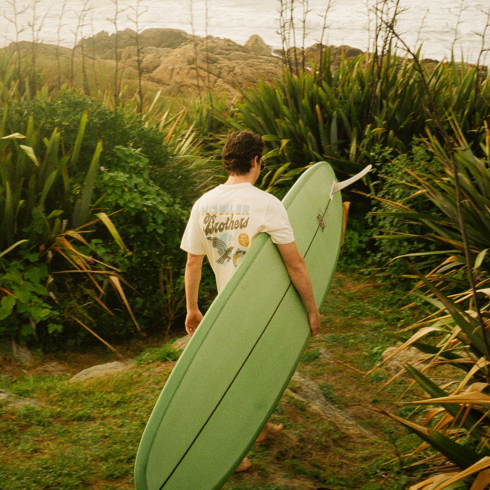 A man carrying a light green surfboard walks through a grassy path surrounded by tropical plants toward the ocean, wearing a white HOWLER POCKET graphic t-shirt.
