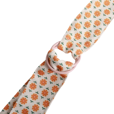 A close-up of a patterned fabric strap with a repeating orange flower and green leaf motif on a cream background, tied with a light pink loop.