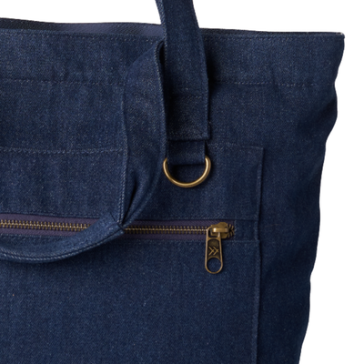 Close-up of a dark navy blue canvas tote bag featuring a front zippered pocket with a brass zipper pull and a matching brass D-ring on the strap.