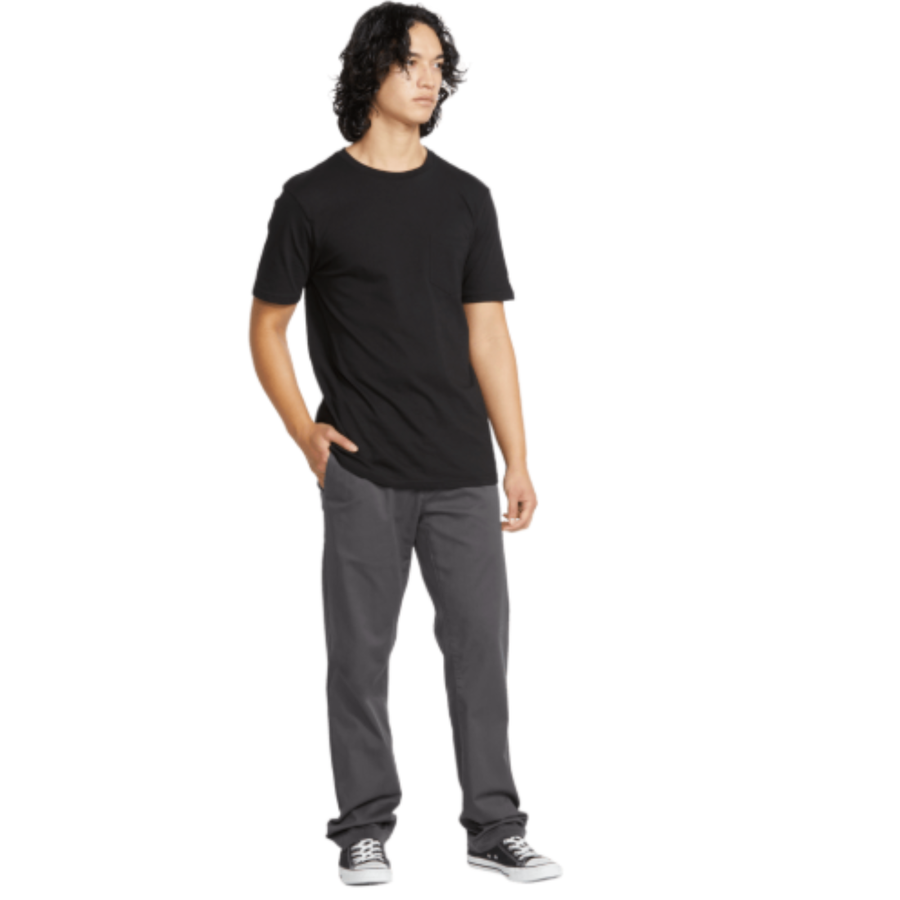 A man wearing a black short-sleeve crewneck t-shirt, dark gray volcom frickin stretch chino pants, and black and white sneakers, standing against a plain white background.