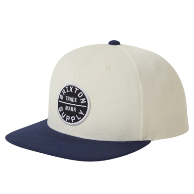 Beige and navy brixton snapback cap with a logo on a white background
