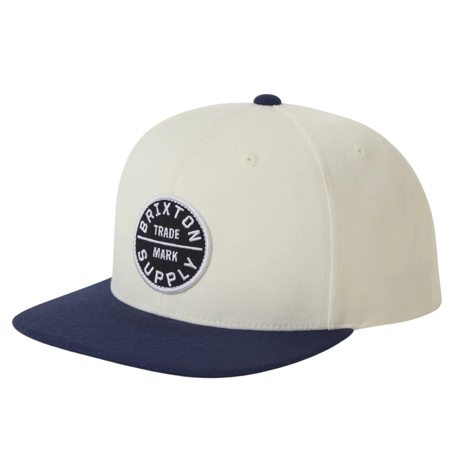Beige and navy brixton snapback cap with a logo on a white background
