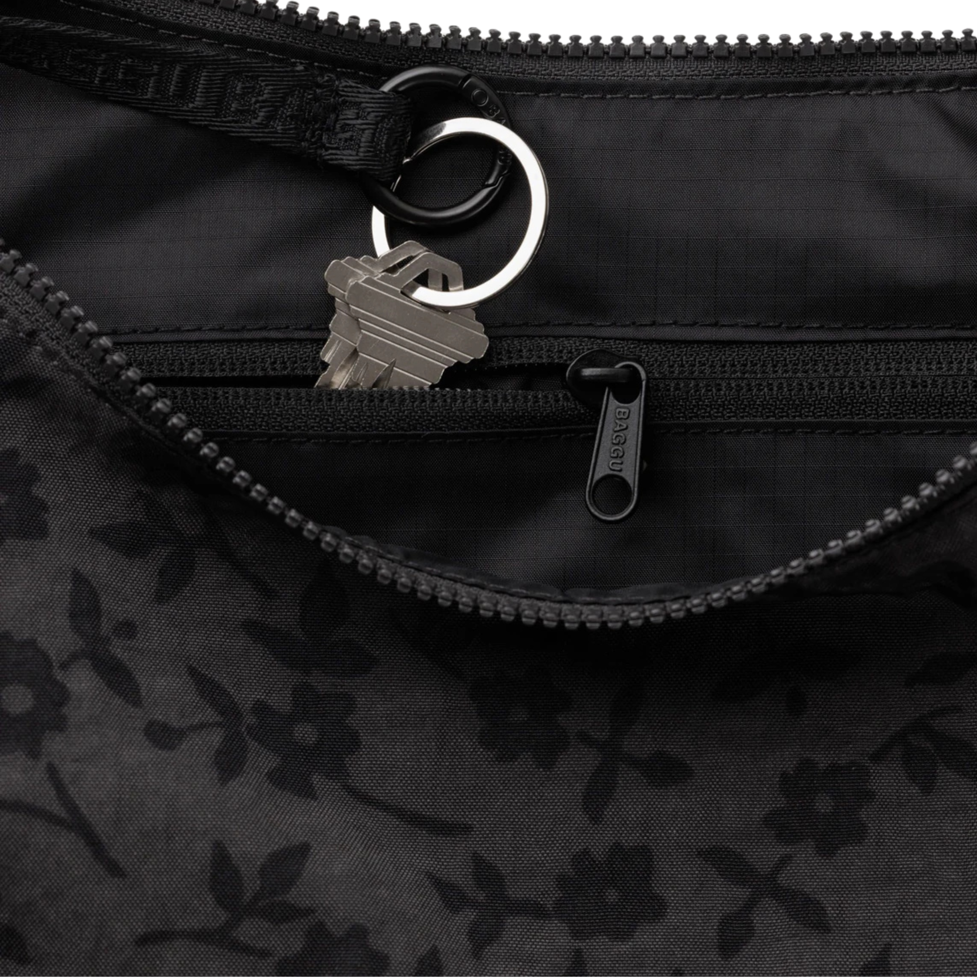 A black crescent-shaped baggu shoulder bag with a subtle tonal floral pattern, featuring a matching black strap with embossed lettering, shown against a plain white background.