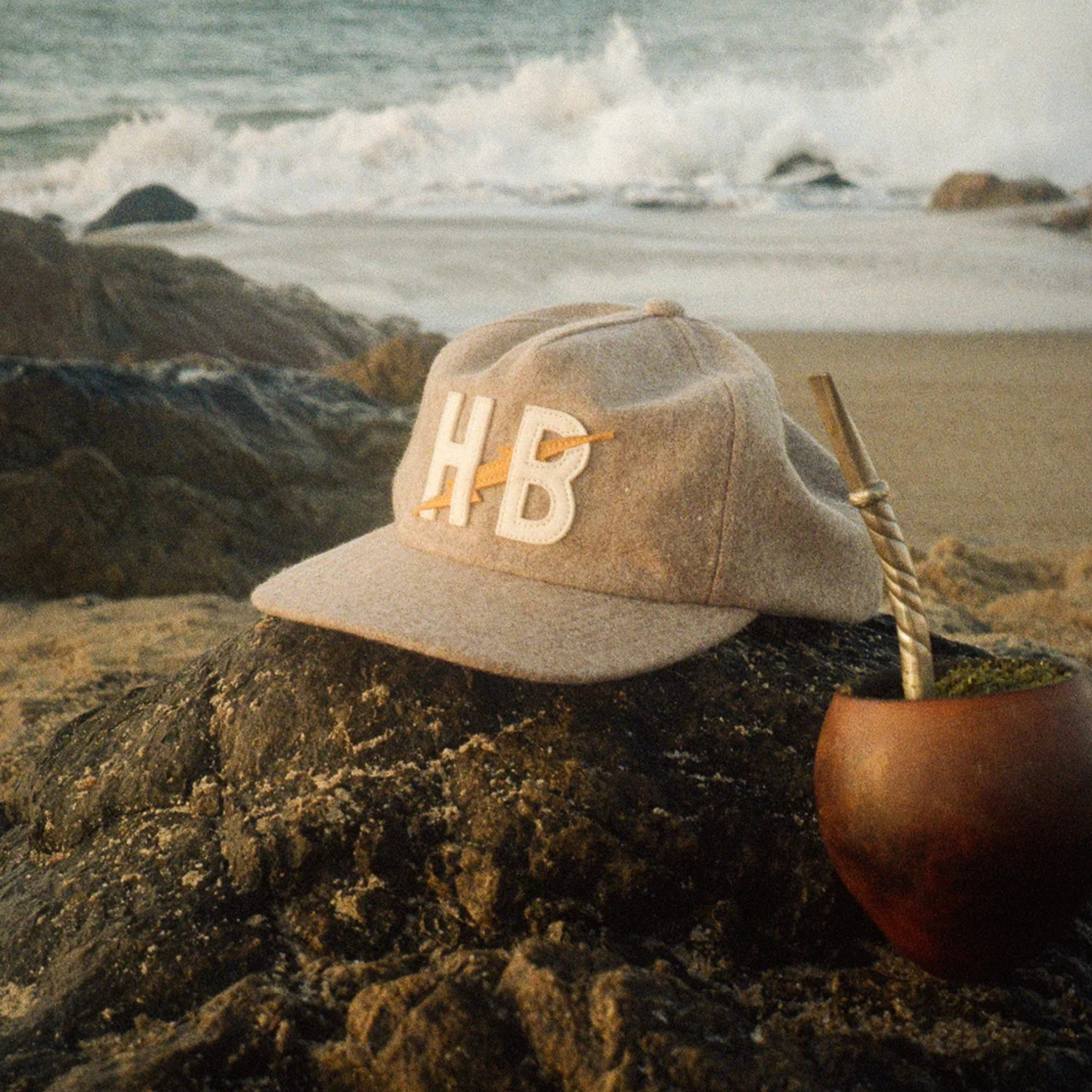 A beige baseball cap with white embroidered letters 'HB' sits on a dark rock at the beach, with ocean waves in the background and a wooden mate cup with a metal straw nearby.