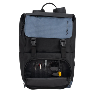 A black and slate blue Volcom backpack shown from the front, with the lower front pocket open to reveal pens, sunglasses, and other small accessories inside. The Volcom logo is printed vertically on the blue flap.