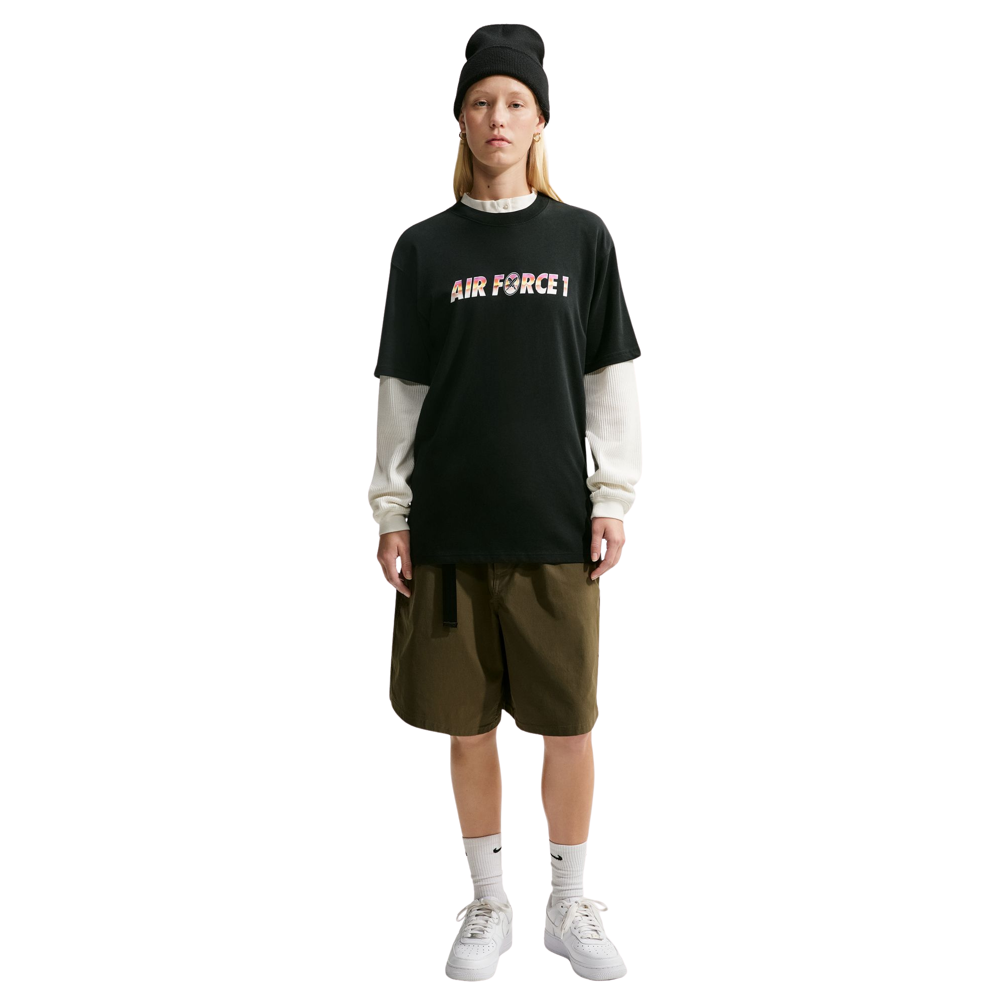 A model stands against a white background wearing a nike sb yuto horigome black t-shirt with 'AIR FORCE 1' printed across the chest in a colorful gradient pattern, layered over a cream long-sleeve shirt. The outfit is completed with olive green shorts, white socks, and white sneakers, and a black beanie.