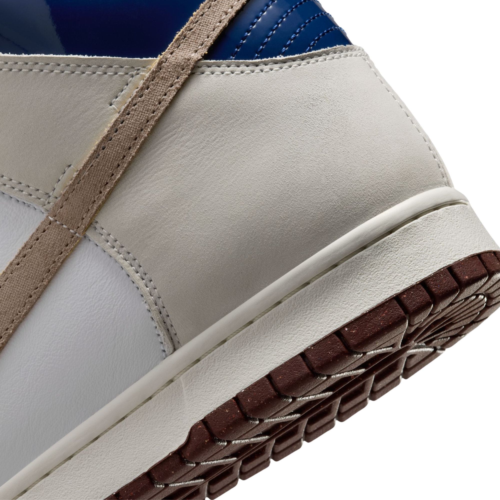 Close-up of a A Nike SB X Beyond Skate Shop  dunk high sneaker showing the rear quarter and midsole, with an off-white leather upper, a tan strap detail, a dark blue collar, and a brown treaded outsole.