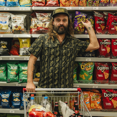 beared man in a grocery store wearing a Holwer Brothers H Bar B Snapshirt grampa's curtains in a black with a gold floral design on a white background grabbing a bag of takis