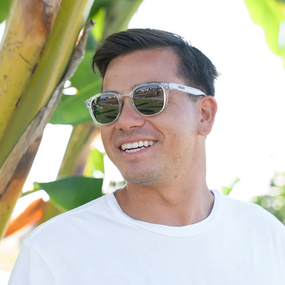 A smiling man wearing clear-framed sunglasses with mirrored lenses and a white t-shirt, photographed outdoors with bright sunlight and blurred green foliage in the background.