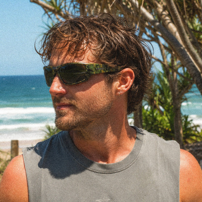 A man outdoors by the beach wearing dark sunglasses with a green camouflage pattern on the arms and frame, along with a gray sleeveless shirt.