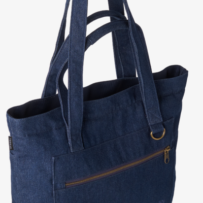 A dark blue denim tote bag with dual shoulder straps, a front zippered pocket, and a brass-tone metal ring detail, shown against a plain white background.