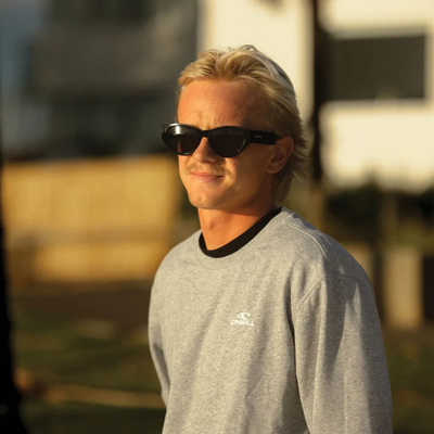 A man wearing black sunglasses and a gray crewneck sweatshirt with a black collar, photographed outdoors in warm golden hour light. The brand name 'O'Neill' is visible on the chest of the sweatshirt.