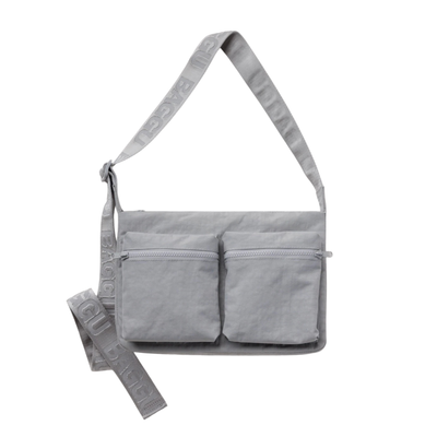 A light gray fabric baggu cargo crossbody shoulder bag with two front zippered pockets and a wide adjustable strap featuring embossed text, shown on a plain white background.