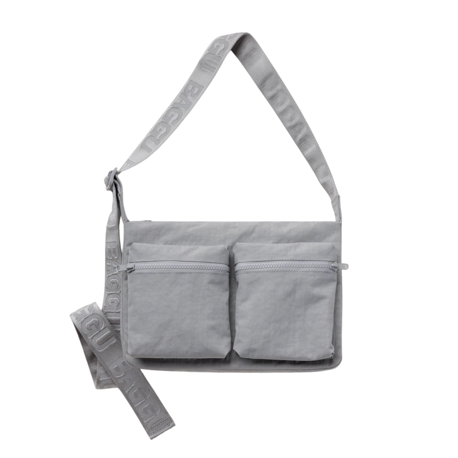 A light gray fabric baggu cargo crossbody shoulder bag with two front zippered pockets and a wide adjustable strap featuring embossed text, shown on a plain white background.
