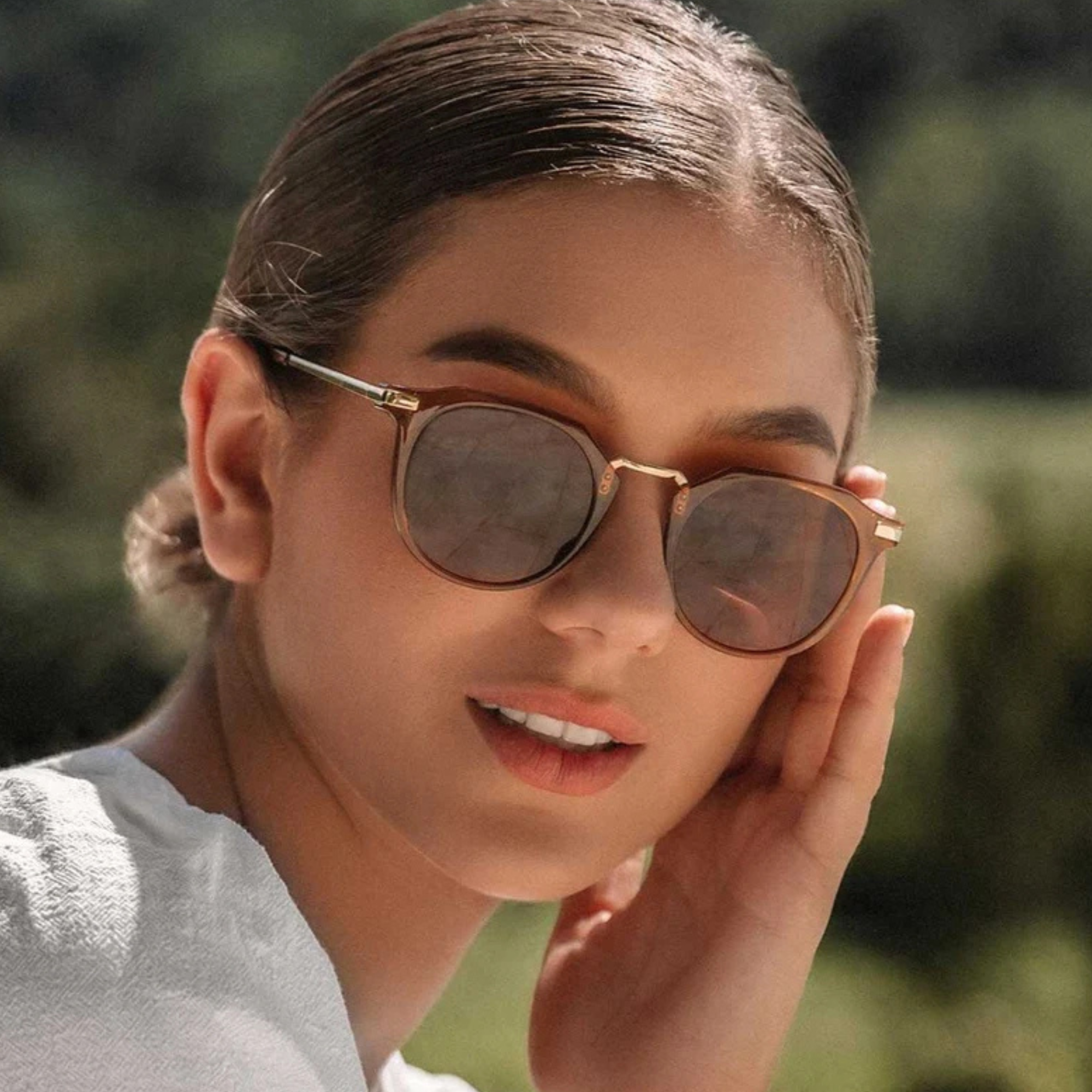 A close-up lifestyle shot of a woman wearing stylish round sunglasses with a bronze metal frame and dark lenses, set against a soft-focus green outdoor background.