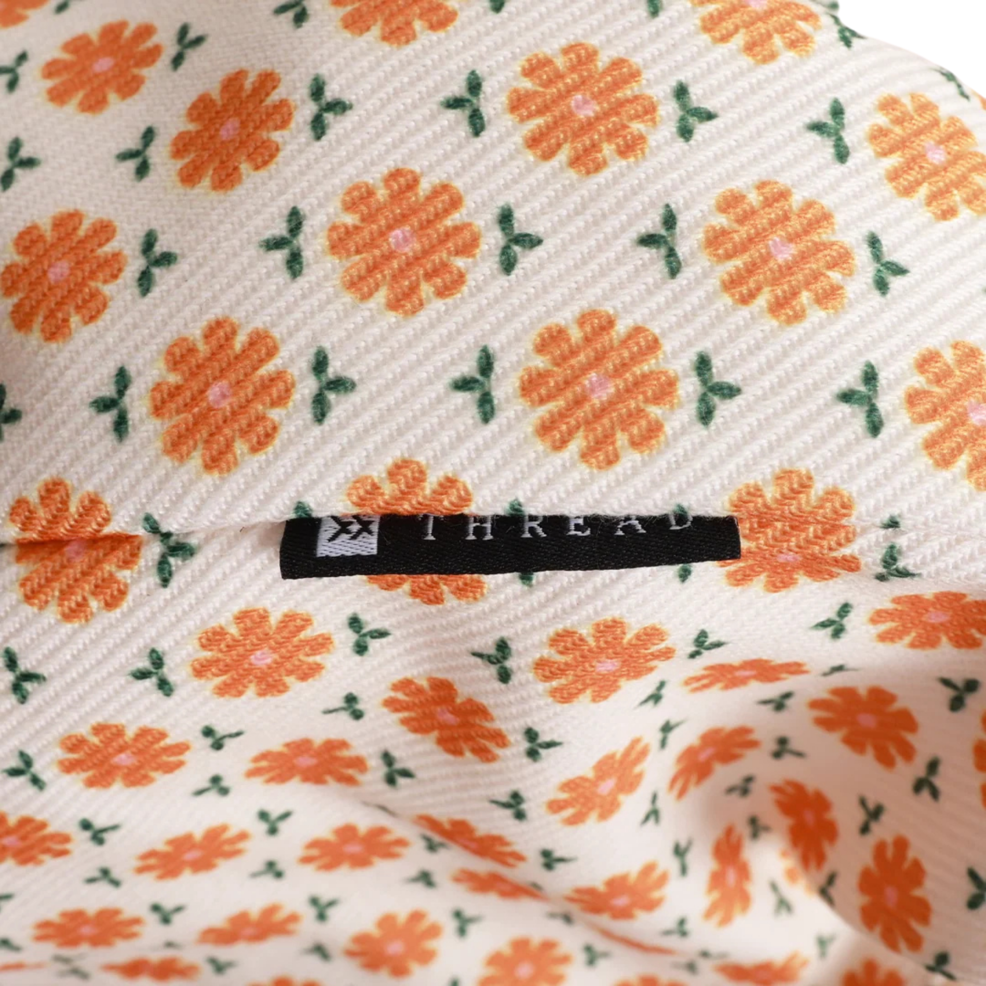 A close-up of a cream-colored woven fabric with a repeating pattern of orange flowers and green leaves, featuring a black fabric brand label sewn into the material. The brand name 'THREAD' is printed on the label.