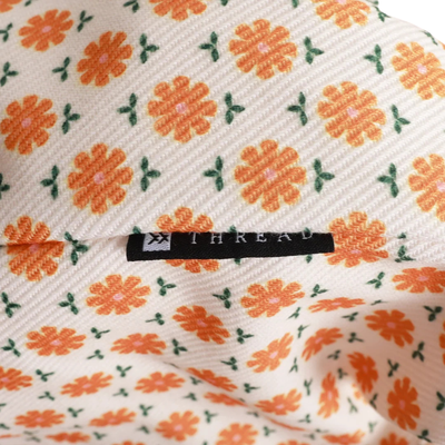 A close-up of a cream-colored woven fabric with a repeating pattern of orange flowers and green leaves, featuring a black fabric brand label sewn into the material. The brand name 'THREAD' is printed on the label.