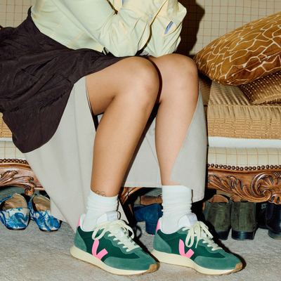 A person seated indoors wearing green low-top veja sneakers with bright pink side stripes, white socks, and a light beige skirt. The setting includes a patterned cushion, wooden furniture, and other shoes on the floor.