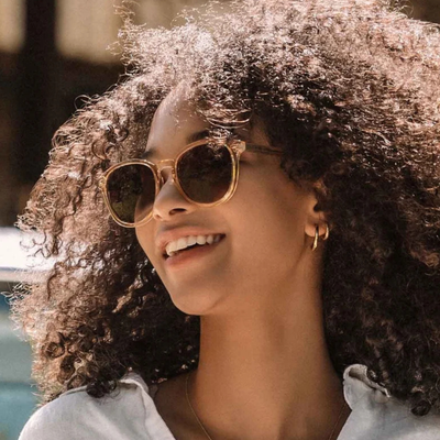 A close-up lifestyle shot of a smiling woman with voluminous curly hair wearing gold-framed carve usa eyewear oslo sunglasses with dark lenses.