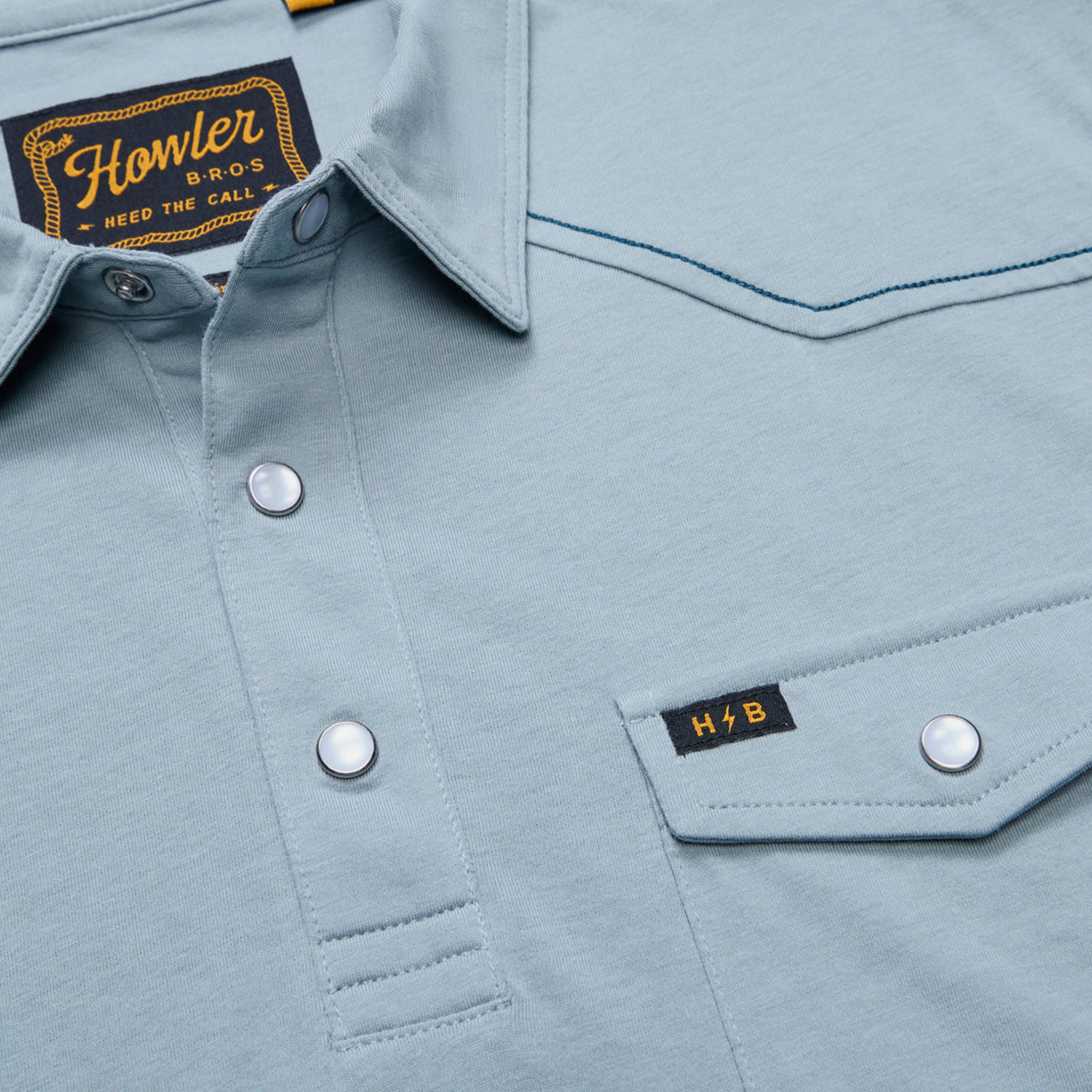 close up of A light blue short-sleeve Howler brothers ranchero polo shirt with western-style stitching, a two-button placket, and a chest pocket