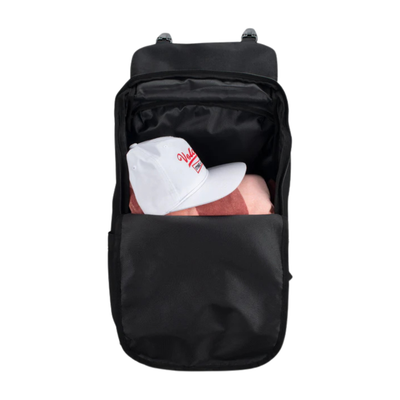 A black backpack shown from the front with its top zip compartment open, revealing a white baseball cap with red lettering and a pink item inside. The backpack is photographed on a solid white background.