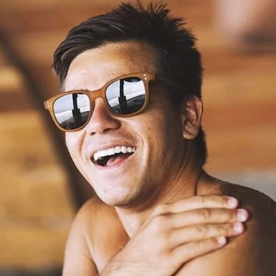 A smiling man wearing brown wood-framed sunglasses with mirrored lenses, shown in a close-up portrait against a warm wooden background.