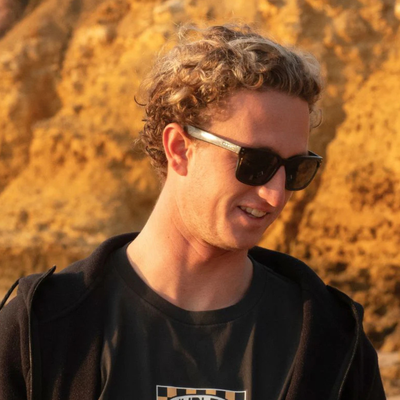 A man with curly light brown hair wearing black sunglasses with silver temples and a black t-shirt, set against a warm, sandy, rocky background.