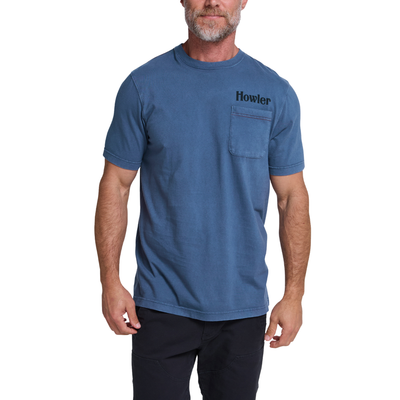 A man wearing a blue short-sleeve crewneck t-shirt with a chest pocket and the brand name 'Howler' printed in black on the left chest, paired with black pants against a white background.