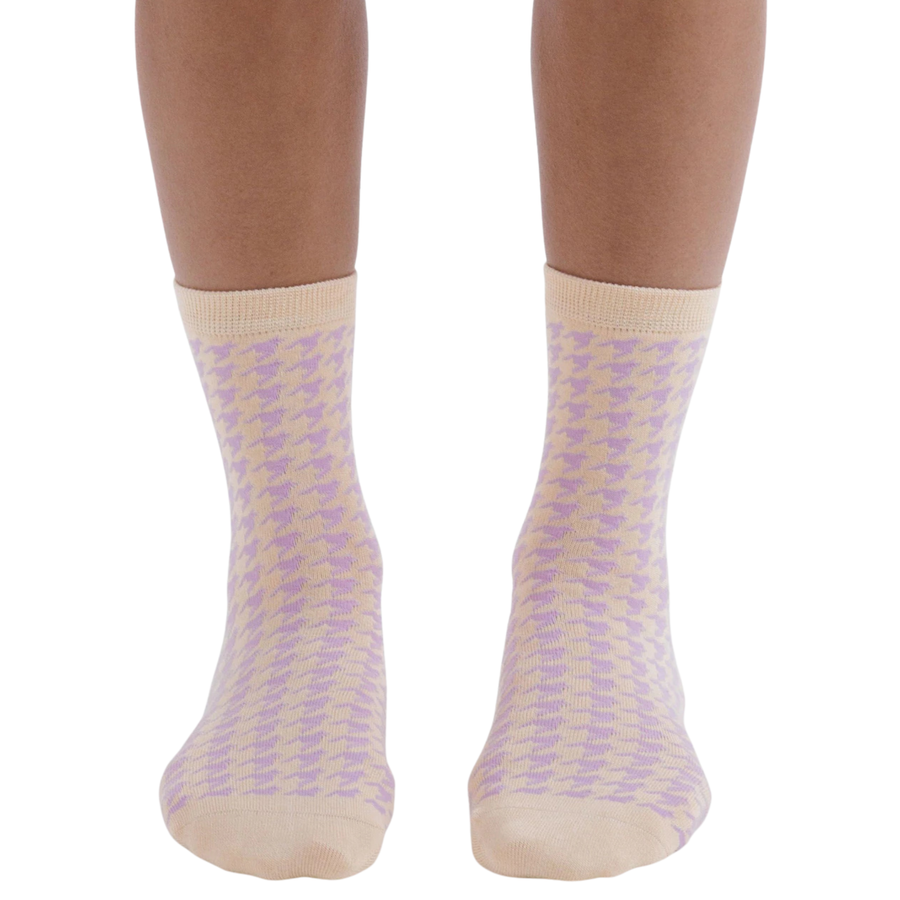 A pair of light pink and lavender houndstooth patterned baggu crew socks with beige ribbed cuffs and toes, featuring the brand name 'BAGGU' printed vertically in olive green on the sole.