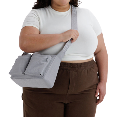 A person wearing a white crop top and brown sweatpants carries A light gray fabric baggu cargo crossbody shoulder bag with two front zippered pockets and a wide adjustable strap featuring embossed text, shown on a plain white background..