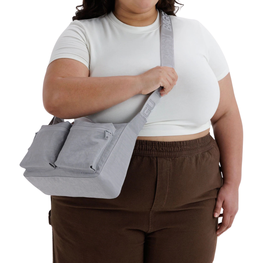 A light gray fabric baggu cargo crossbody shoulder bag with two front zippered pockets and a wide adjustable strap featuring embossed text, shown on a plain white background.