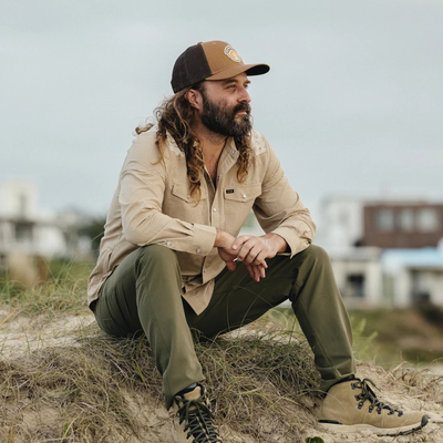 A man with long curly hair and a beard sits outdoors on dry grass, wearing a tan and brown trucker hat, a beige button-up shirt, olive green pants, and tan lace-up boots.
