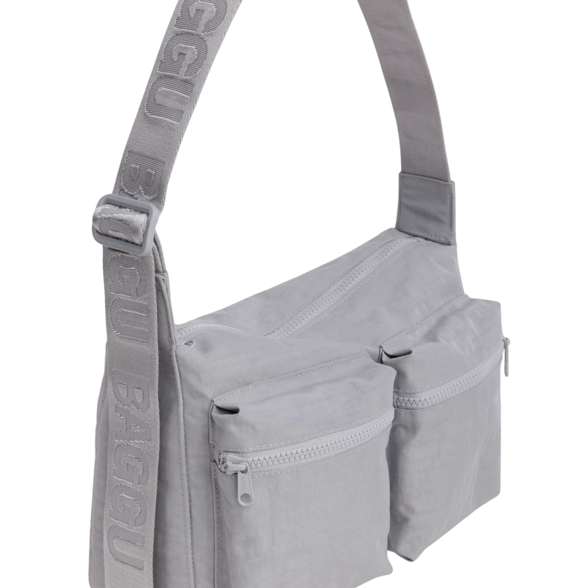 A light gray fabric baggu cargo crossbody shoulder bag with two front zippered pockets and a wide adjustable strap featuring embossed text, shown on a plain white background.